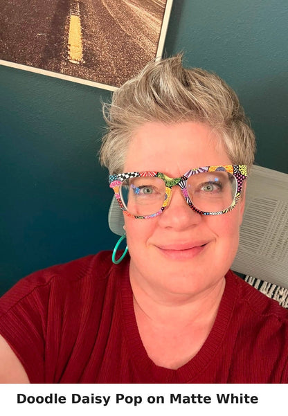 A close-up of a person wearing colorful patterned eyeglasses with a multicolor daisy print frame, paired with a red top and turquoise earring, against a dark teal wall background.